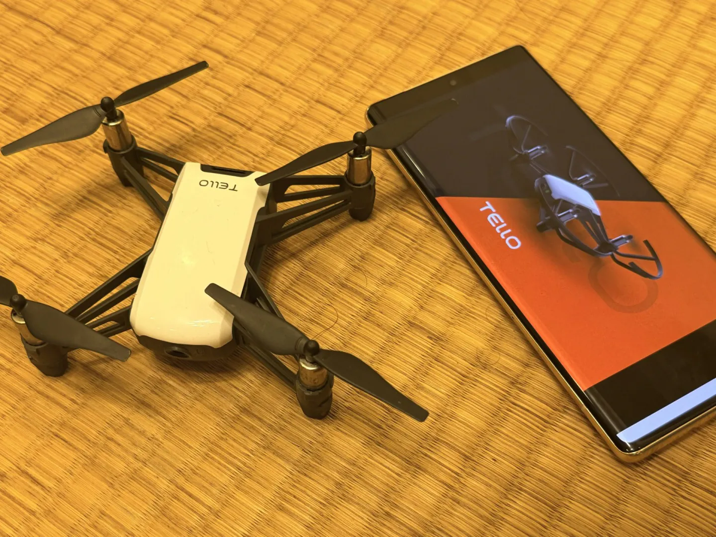 木桌上的白色 Ryze Tello 無人機與 Android 手機，手機螢幕顯示 Tello App 的啟動畫面。